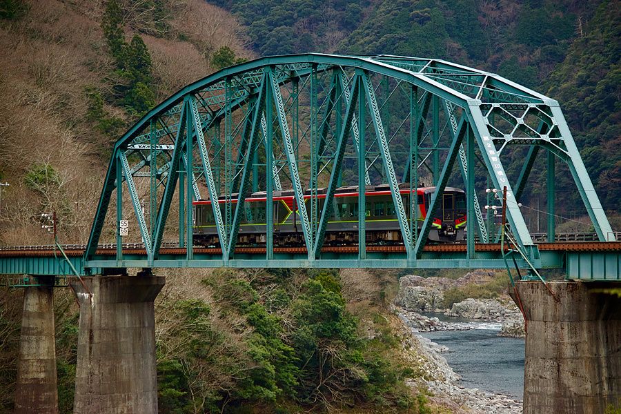 Un treno a strisce rosse e nere attraversa un ponte a traliccio verde sopra una gola in Giappone.