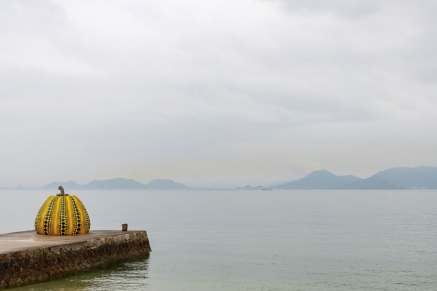 Scultura iconica di una zucca gialla a pois posizionata su un molo di cemento che si affaccia sul mare con colline sullo s...