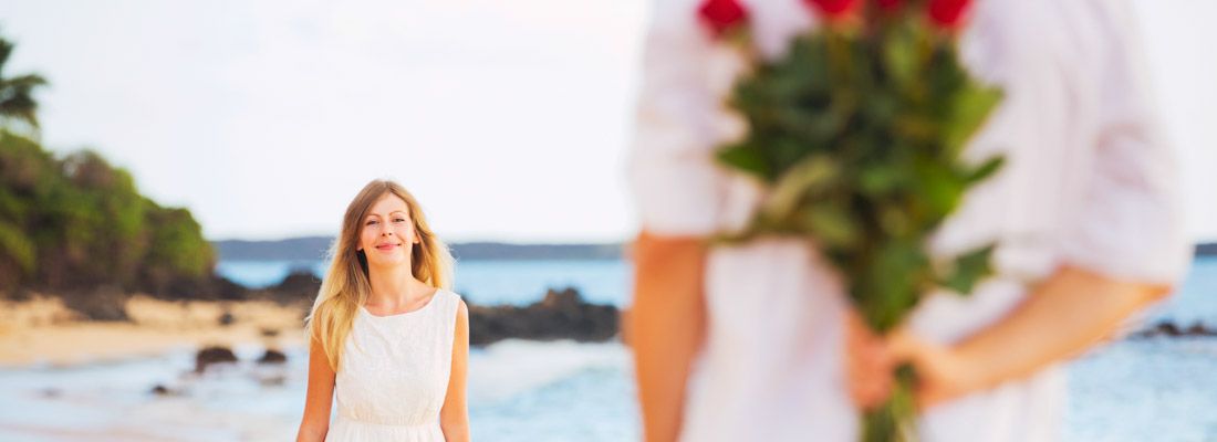 Un ragazzo in spiaggia, con delle rose dietro la schiena, fa una sorpresa ad una ragazza.