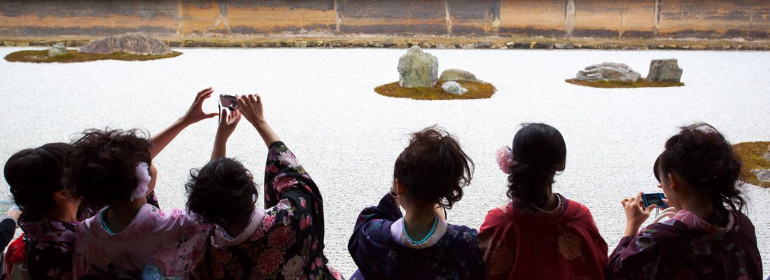 Ragazze in kimono sedute ammirano e fotografano il santuario Ryoanji.