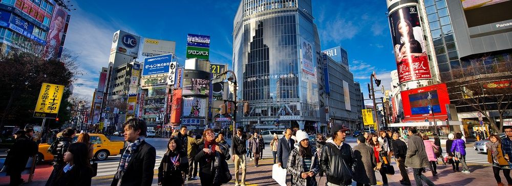 Persone attraversano l'incrocio di Shibuya a Tokyo, circondato da edifici e schermi luminosi.