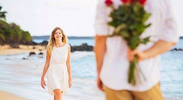 Un ragazzo in spiaggia, con delle rose dietro la schiena, fa una sorpresa ad una ragazza.