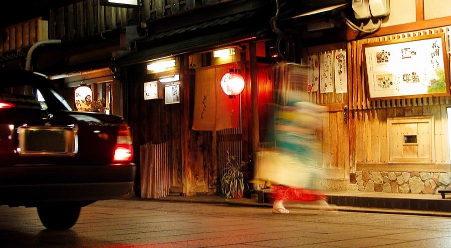 Strada notturna nel quartiere di Gion, Kyoto, con lanterne e figure in movimento.