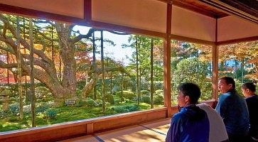 Persone osservano un giardino giapponese attraverso grandi finestre in una casa tradizionale.